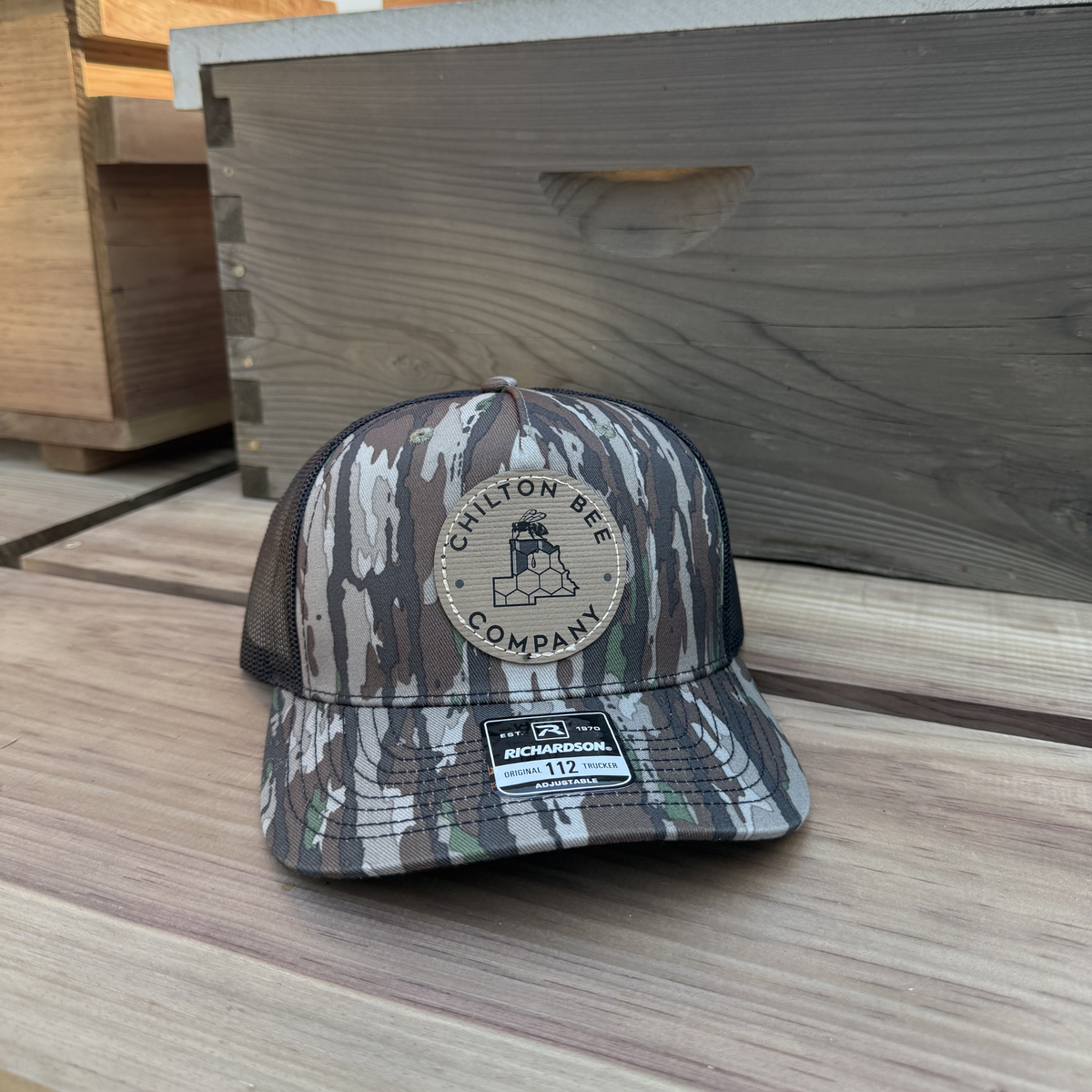Chilton Bee Company Richardson 112 trucker hat in black and gray camo with black mesh back and round leather logo patch, displayed on a wooden surface next to a beehive box