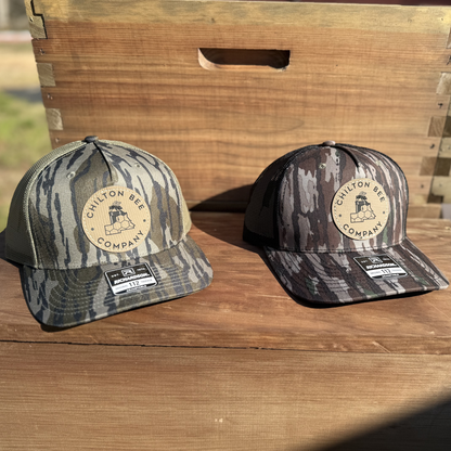  Two Chilton Bee Company Richardson 112 trucker hats in mossy oak and black camo patterns with leather logo patches, displayed in front of a wooden beehive box