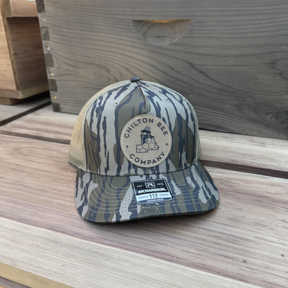 Chilton Bee Company Richardson 112 trucker hat in mossy oak camo with tan mesh back and round leather logo patch, resting on a wooden surface near a beehive